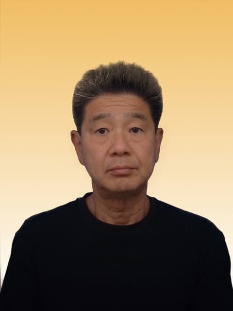 代表・新飯田啓二の顔写真(黒のトップス、明るいベージュ調の背景)